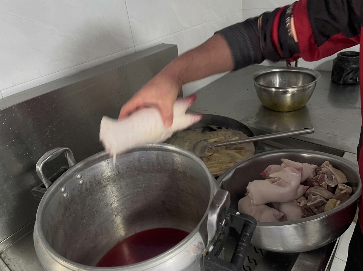 Eine Hand legt Schweinefüße in einen großen Topf mit dunkler Schmorflüssigkeit.