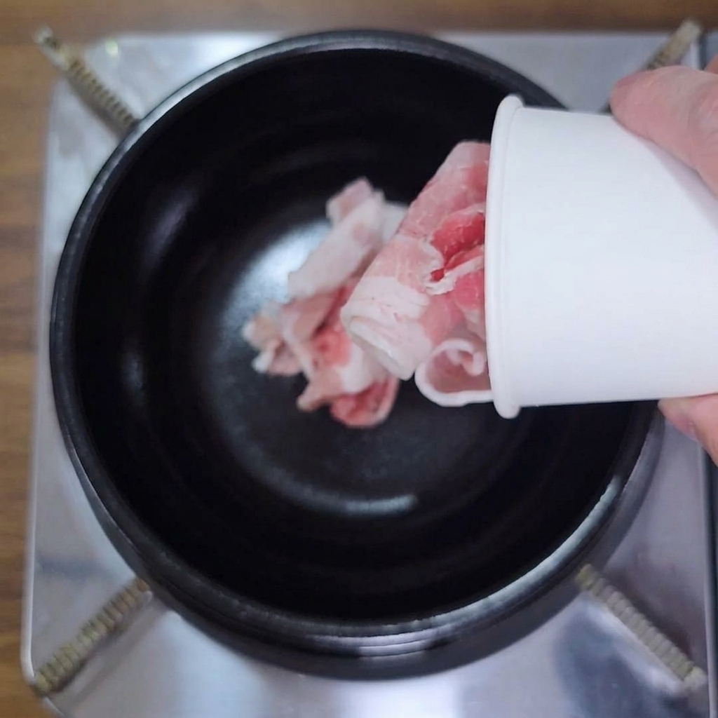 Eine Hand schüttet gerollten, dünn geschnittenen rohen Schweinebauch aus einem weißen Pappbecher in einen schwarzen Topf.