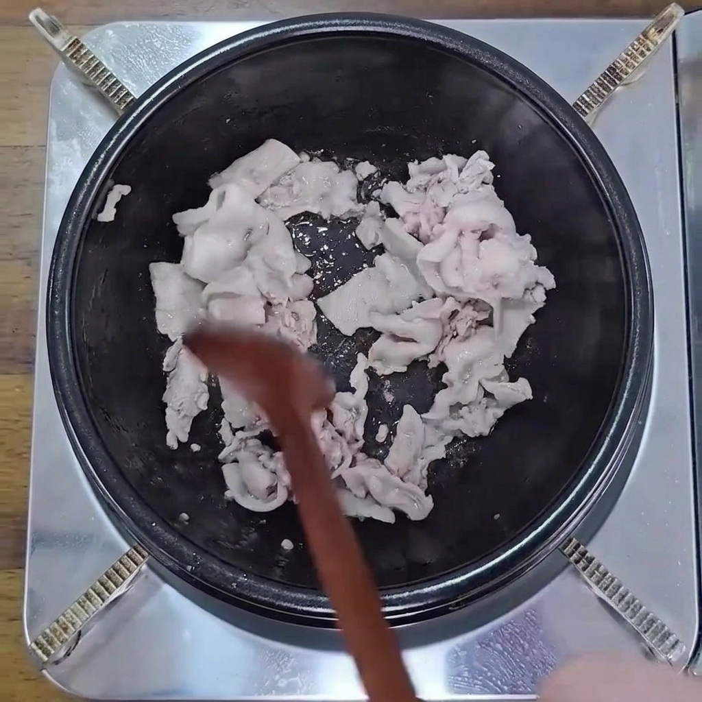Dünn geschnittener Schweinebauch wird in einem schwarzen Topf mit einem Holzlöffel angebraten, bis er leicht gebräunt ist.