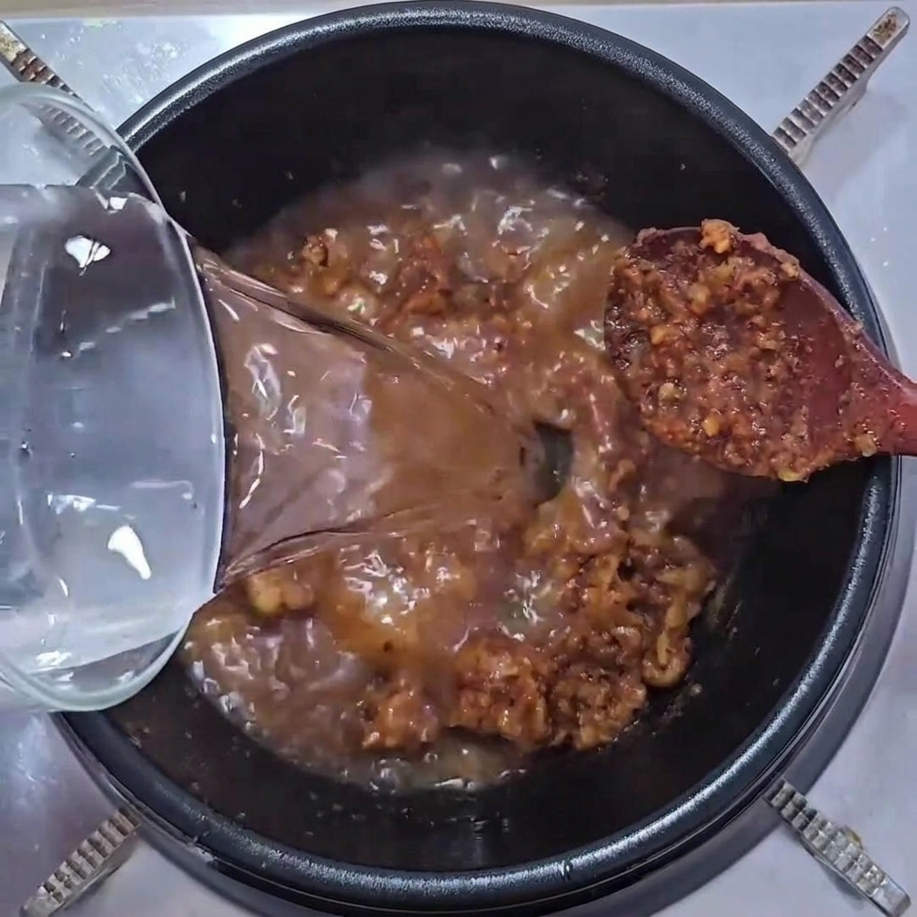 Klares Wasser wird aus einem Glasgefäß in einen Topf mit einer brutzelnden Mischung aus Schweinefleisch und Sojabohnenpaste gegossen.