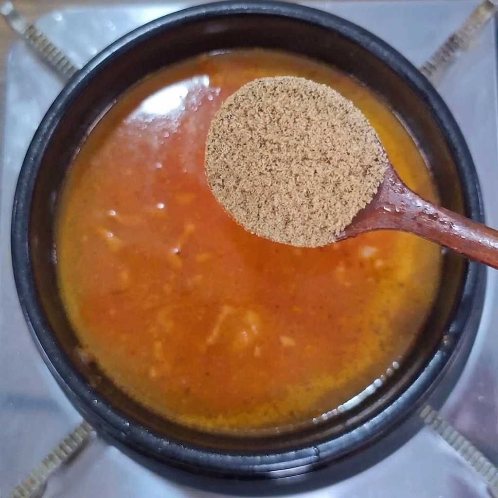 Ein Holzlöffel mit trockenem Rinderbrühe-Pulver über einem Topf mit sprudelnder orangeroter Brühe.