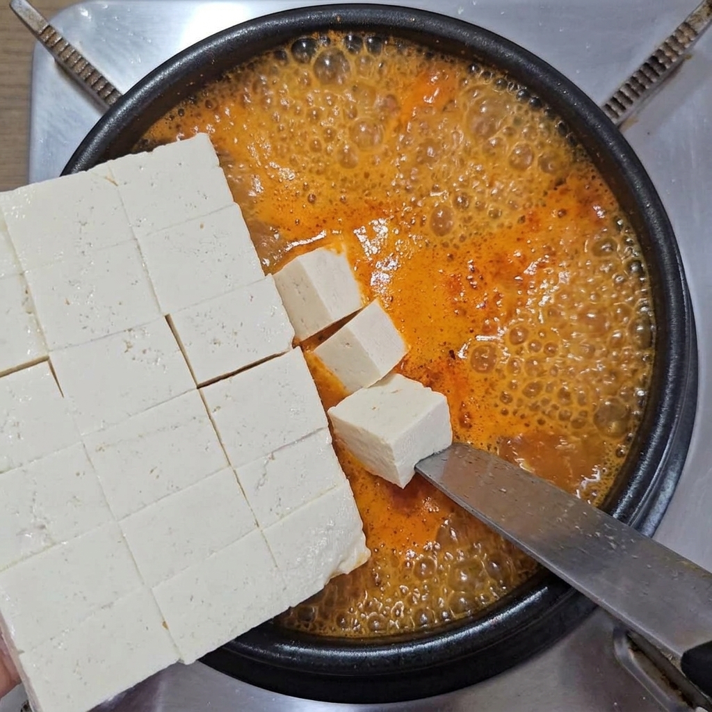 Gewürfelter weicher weißer Tofu wird vorsichtig von einem großen Messer in einen Topf mit brodelndem orangerotem Eintopf geschoben.