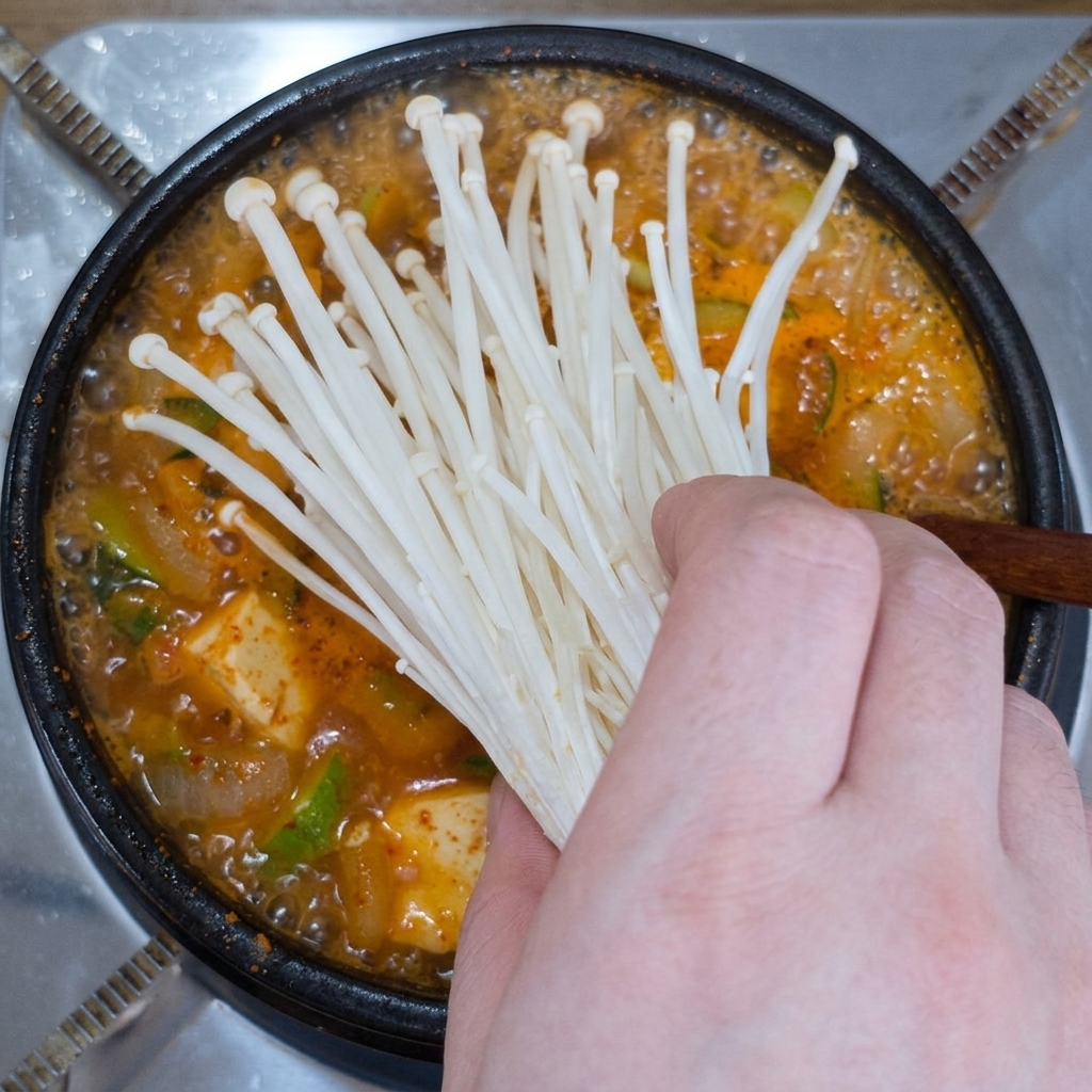 Eine Hand legt ein Bund frische weiße Enoki-Pilze in einen Topf mit kochendem Sojabohnenpasten-Eintopf.