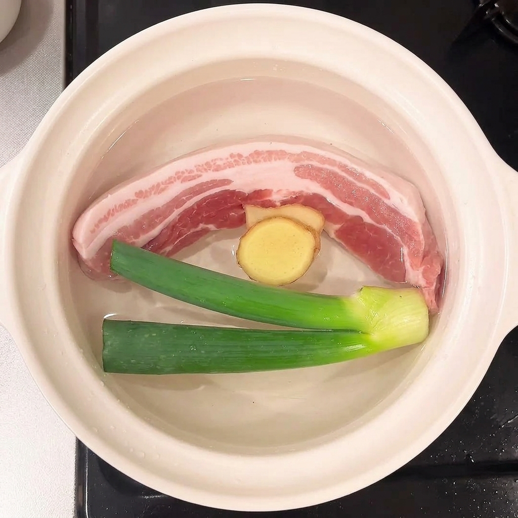Frühlingszwiebeln und Ingwerscheiben schwimmen neben dem Schweinebauch im Wasser.