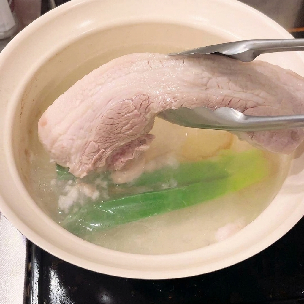 Eine Zange hebt den blanchierten, festen Schweinebauch aus dem Kochwasser.