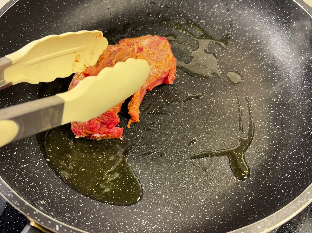 Ein Stück gewürztes Rindfleisch wird in einer heißen Pfanne mit Öl angebraten und mit einer Zange festgehalten.