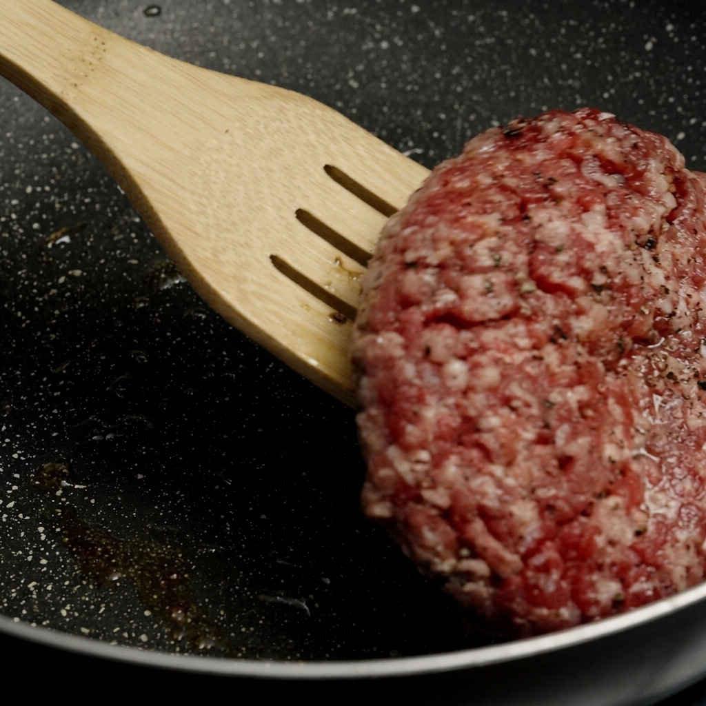 Ein Pfannenwender aus Holz hebt und wendet ein dickes Burger-Patty mit einer schön gebräunten Kruste in einer heißen Pfanne.