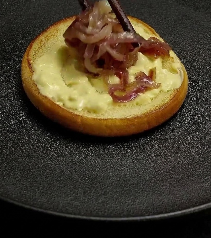 Gebratene rote Zwiebeln werden mit Essstäbchen auf ein Brötchen gelegt, das mit cremiger Burgersauce bestrichen ist.