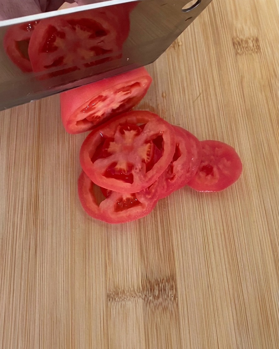 Ein Messer schneidet eine reife, rote Tomate auf einem Holzbrett in dicke Scheiben.