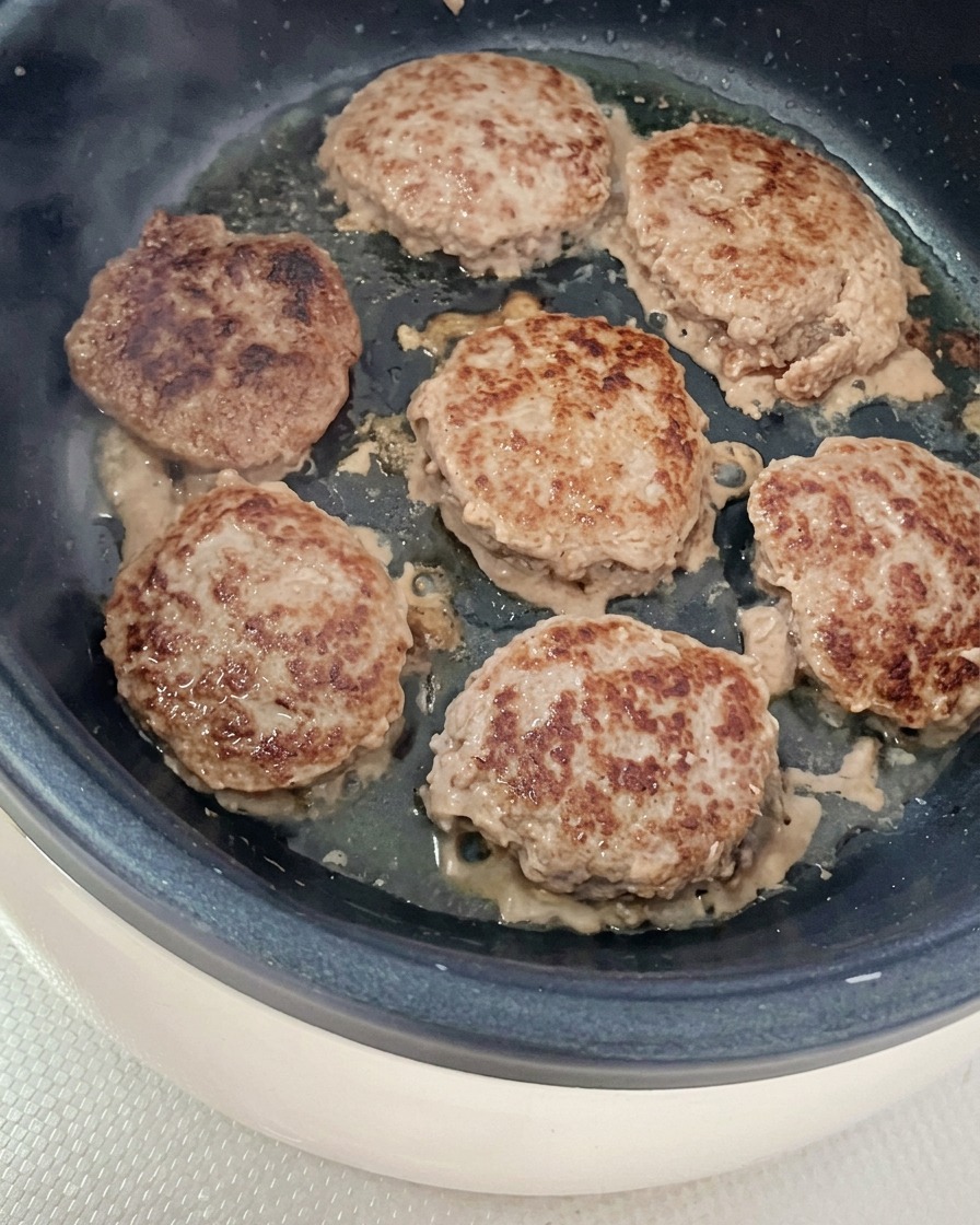 Mehrere runde Rindfleisch-Patties brutzeln und bräunen in einer leicht geölten Pfanne.