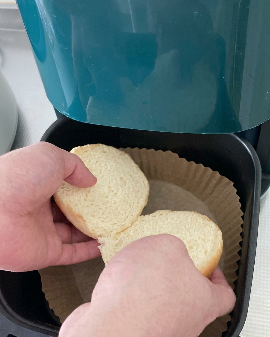 Zwei Hälften eines weißen Burgerbrötchens werden in den mit Backpapier ausgelegten Korb einer Heißluftfritteuse gelegt.