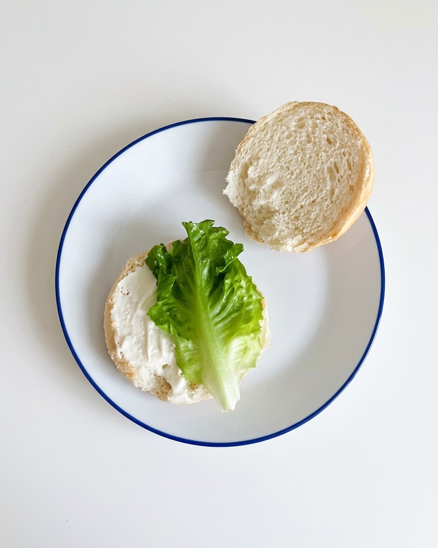 Frischer grüner Salat wird auf die mit Sauce bestrichene untere Hälfte eines Hamburgerbrötchens gelegt.