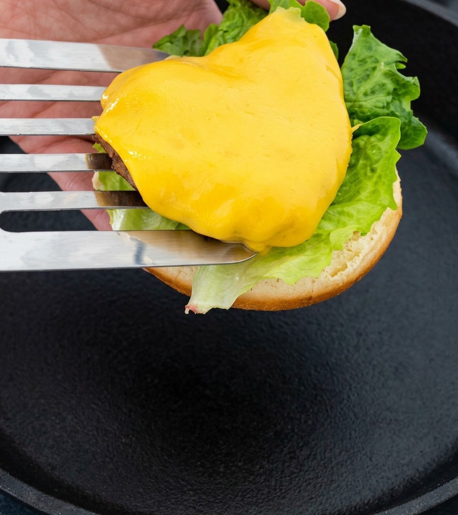 Ein Metall-Pfannenwender hebt ein fleischiges Patty mit Käse an, um es auf das Burgerbrötchen zu legen.