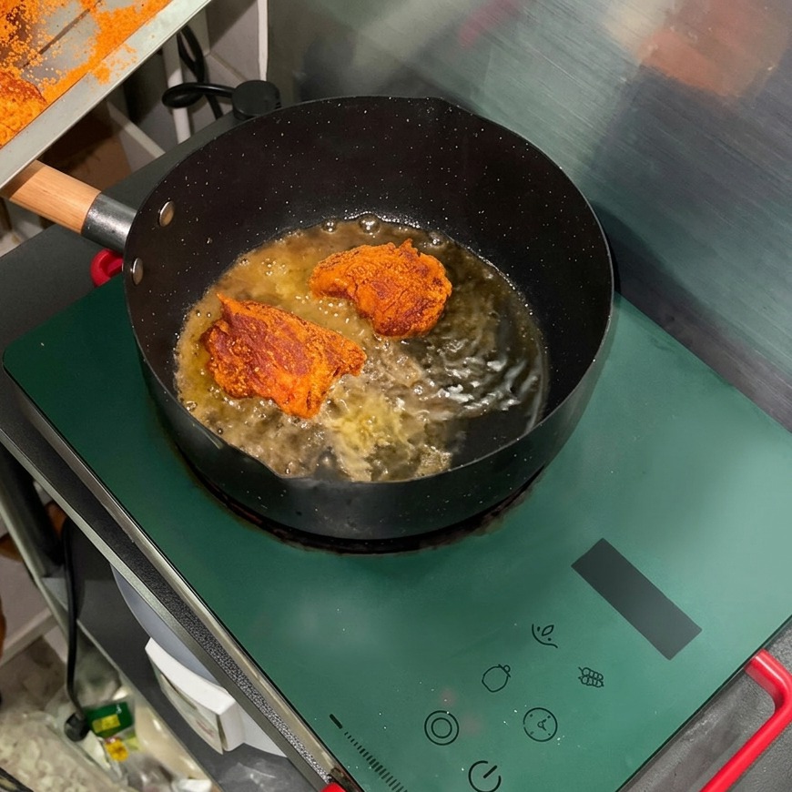 Gewürzte Fleischstücke braten in heißem Öl in einer schwarzen Pfanne auf einem grünen Induktionsherd.
