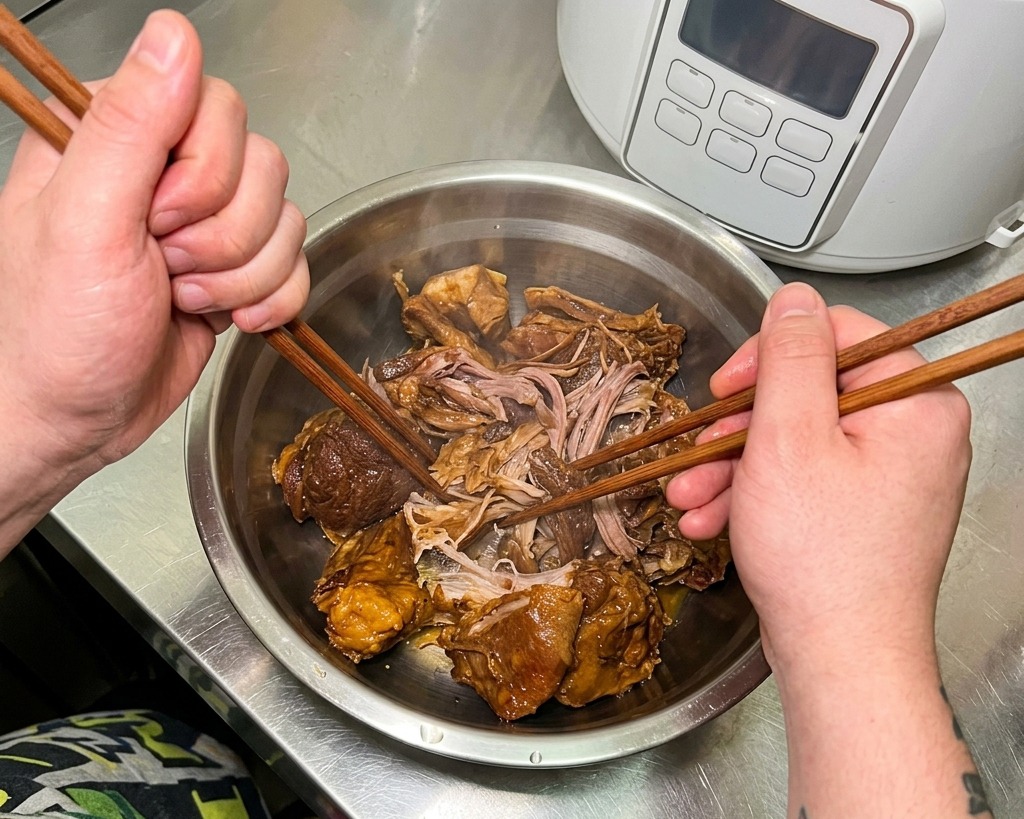 Hände benutzen zwei Essstäbchen, um zartes, geschmortes Fleisch in einer Edelstahlschüssel zu zerteilen.