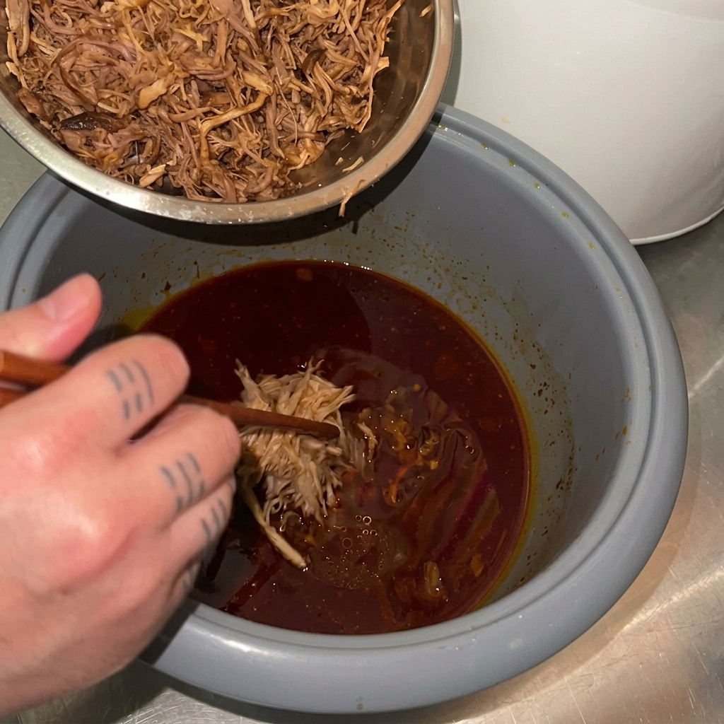 Gezupftes Fleisch wird aus der Schüssel zurück in den Reiskocher zur dunklen Sauce gegeben.