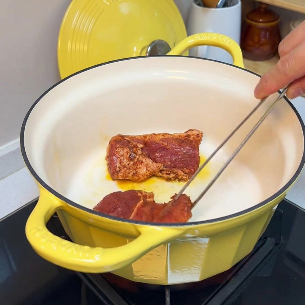 Eine Zange wendet gewürzte Schweinefleischstücke in einem gusseisernen Topf, während sie rundherum anbraten.