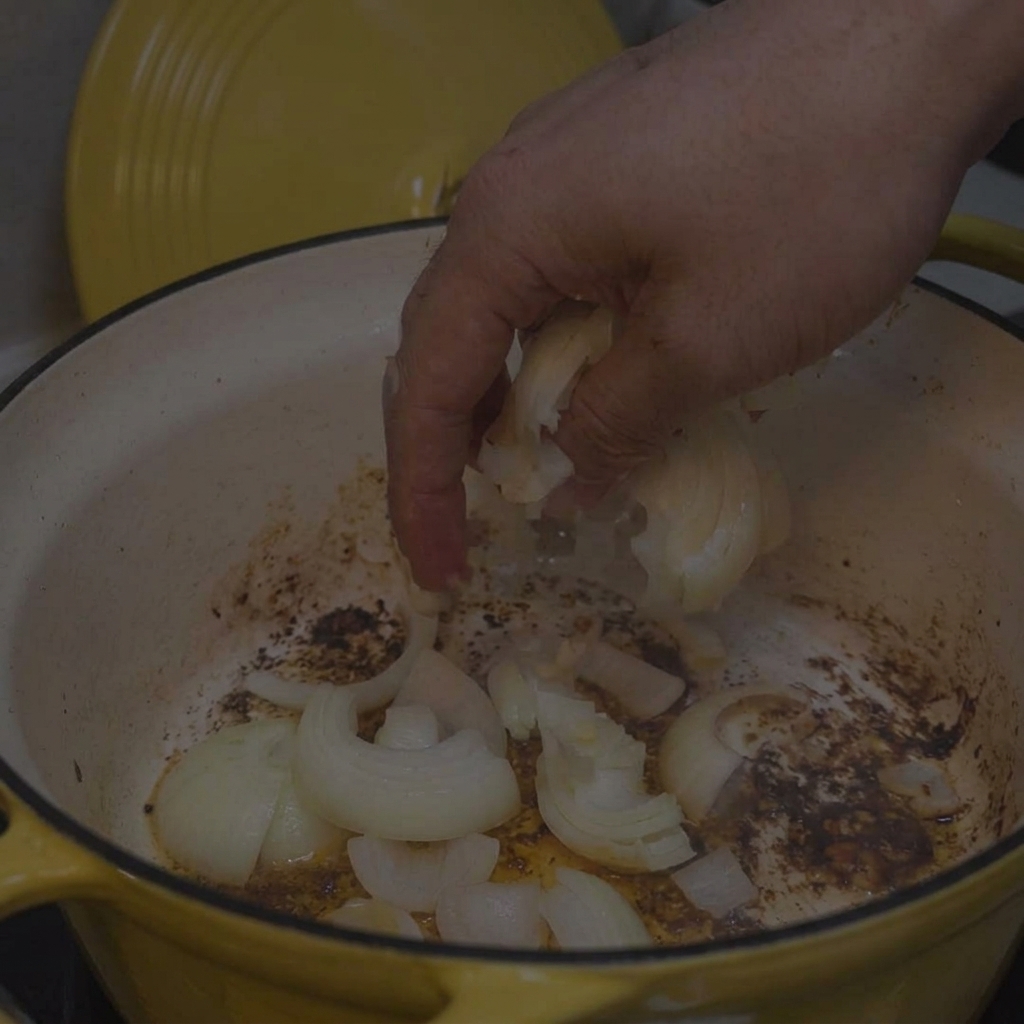 Eine Hand gibt gehackte Zwiebeln in den Topf, in dem sich noch Bratrückstände vom Fleisch befinden.