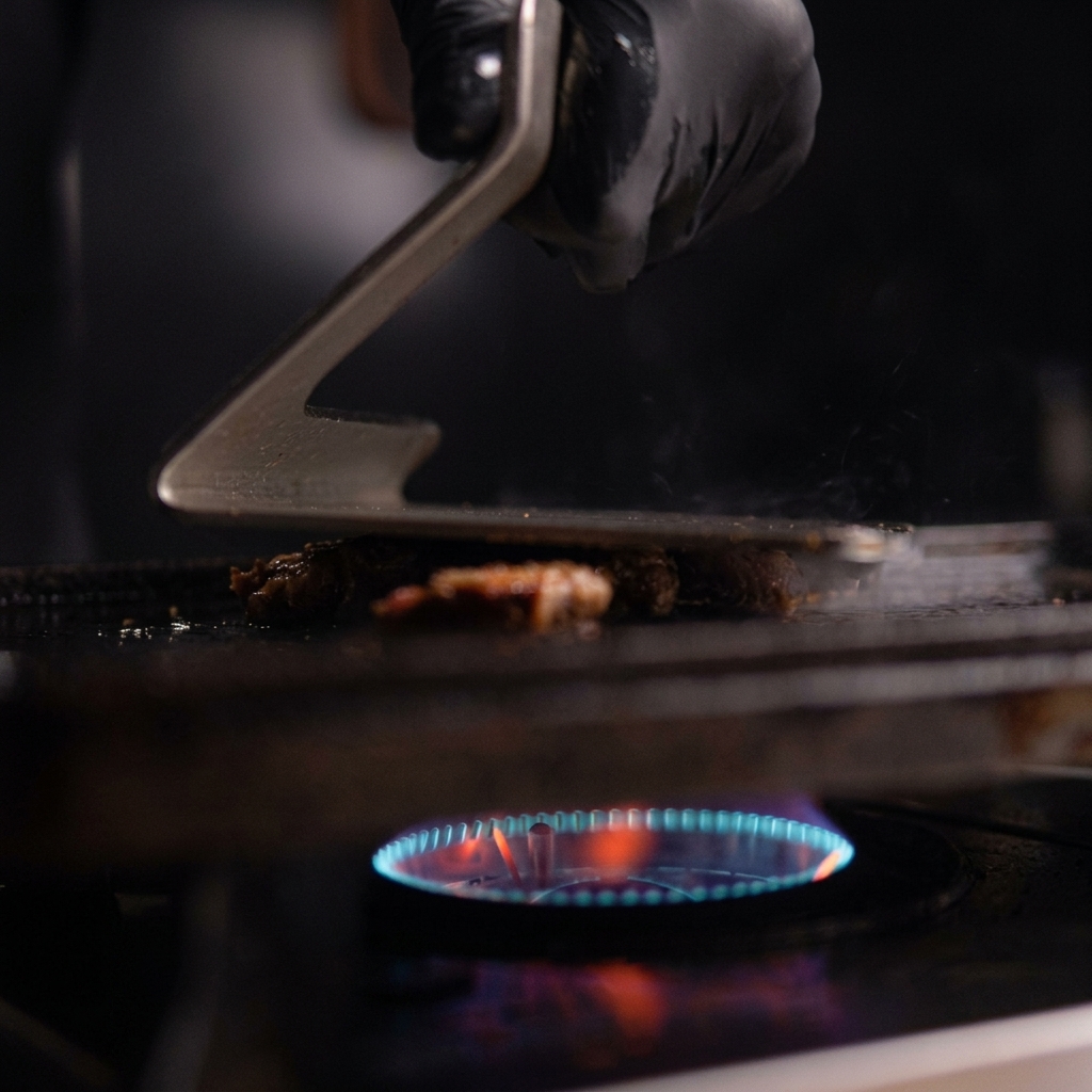 Eine behandschuhte Hand drückt mit einer Grillpresse einen Haufen Pulled Pork auf einer heißen Gusseisenplatte flach.