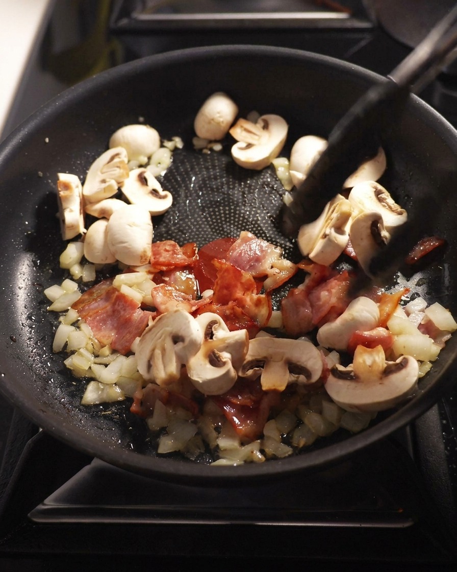 In Scheiben geschnittene Champignons braten zusammen mit Speck, Zwiebeln und Knoblauch in einer Pfanne.