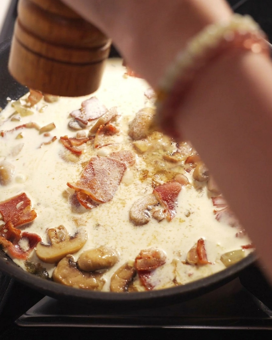 Eine Hand mahlt mit einer Pfeffermühle frischen schwarzen Pfeffer über eine köchelnde Sahnesauce mit Speck und Pilzen.