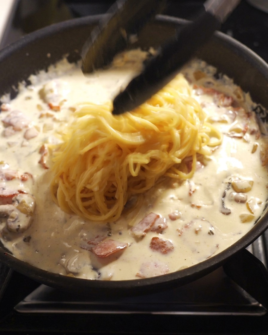Schwarze Küchenzange hebt frisch gekochte Spaghetti in eine Pfanne mit weißer Sahnesauce, Speck und Champignons.