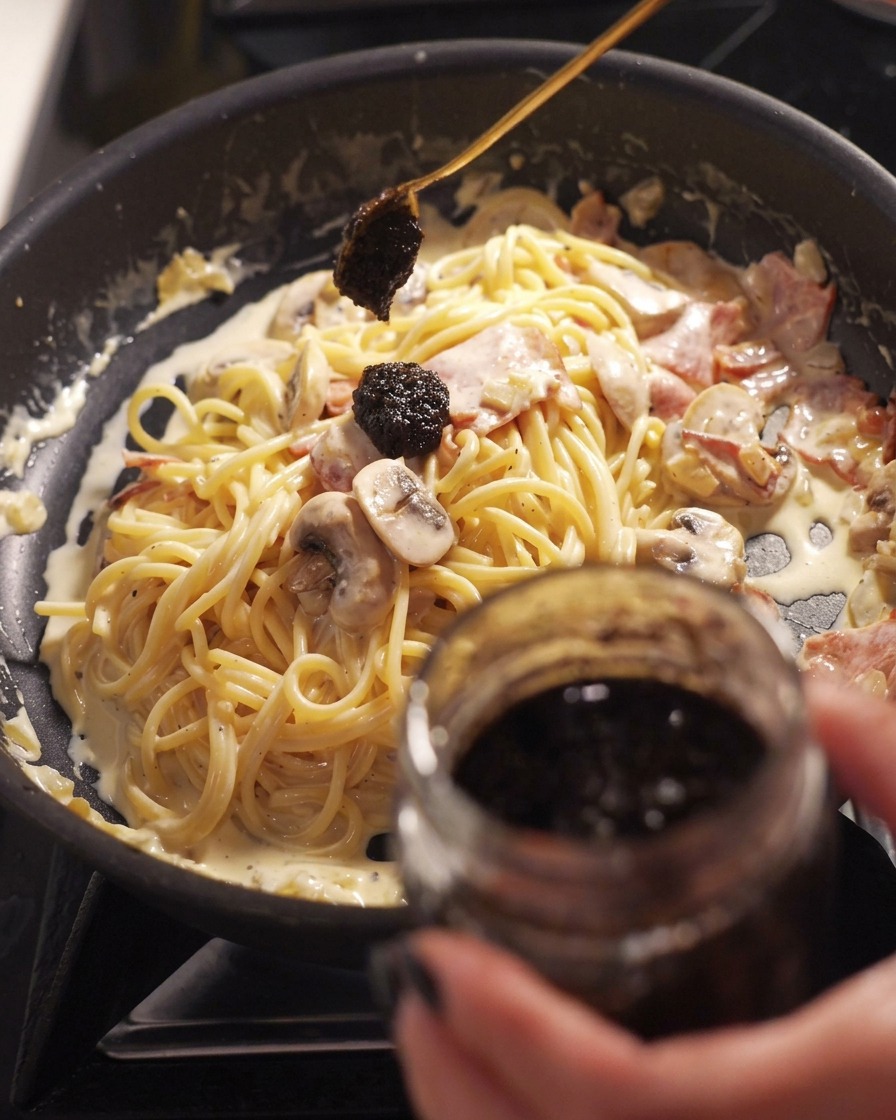 Ein Löffel gibt einen dunklen Klecks schwarze Trüffelpaste in eine Pfanne mit cremigen Spaghetti, Speck und Pilzen.