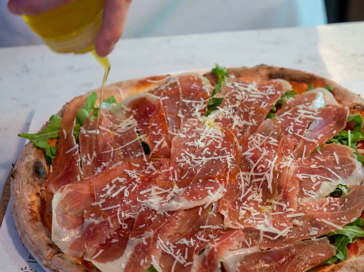 Eine Hand beträufelt eine mit Rucola, Parmaschinken und Käse belegte Pizza mithilfe einer Dosierflasche mit Olivenöl.