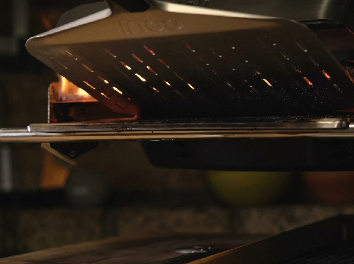 Eine Pizza auf einem Metallschieber wird in einen glühend heißen, tragbaren Pizzaofen geschoben, in dem leuchtend orangefarbene Flammen lodern.