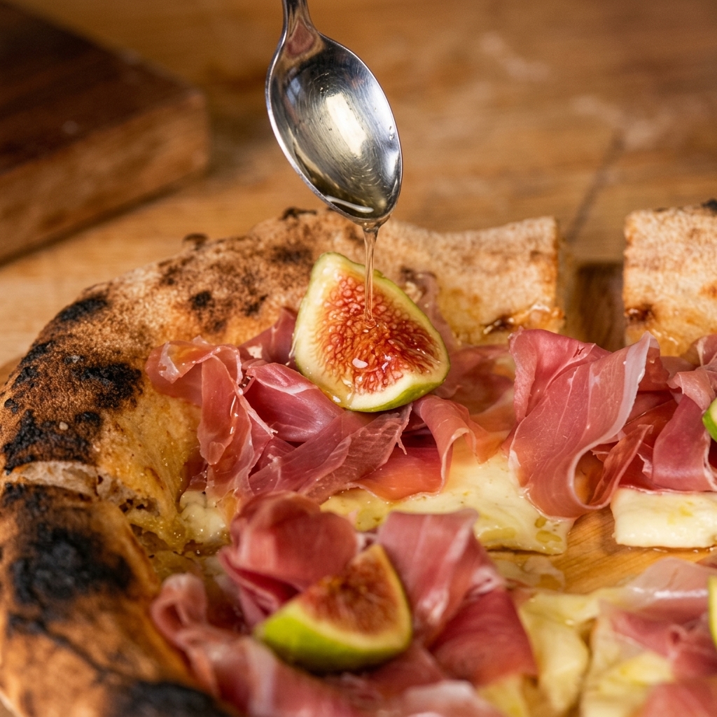 Ein silberner Löffel träufelt klaren Honig über eine rustikale Pizza, die mit geschmolzenem Käse, gefalteten Parmaschinkenscheiben und einer halbierten frischen Feige belegt ist.