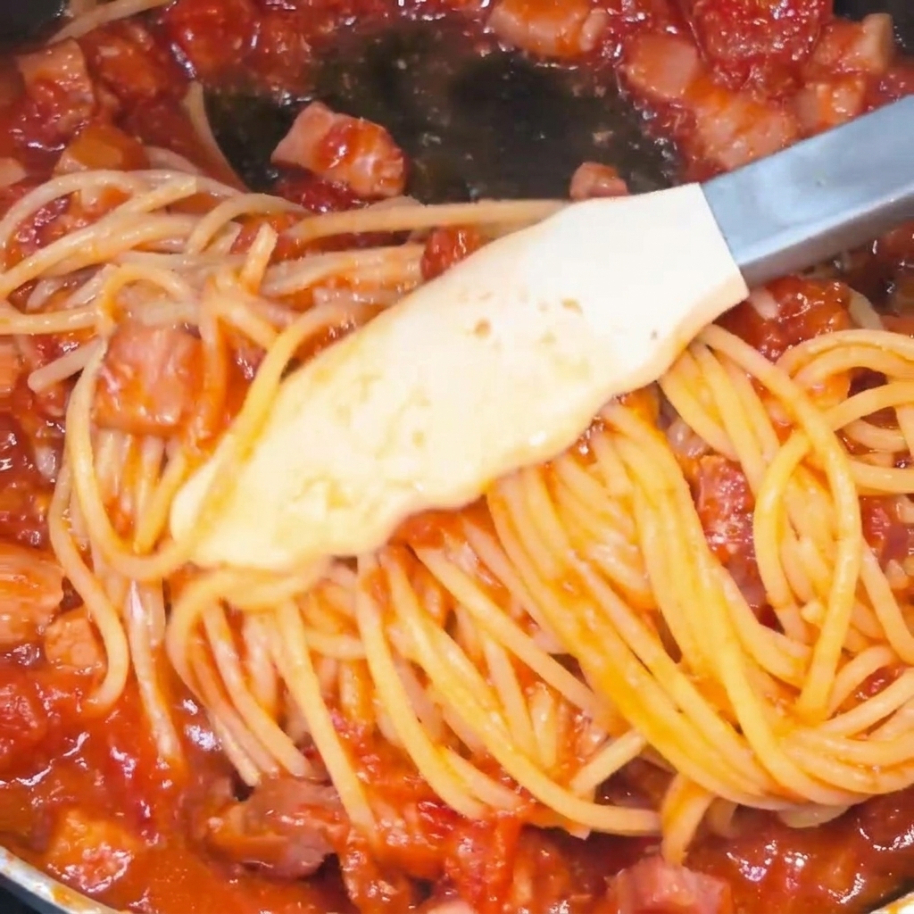 Spaghetti werden mit einer Zange in einer Pfanne geschwenkt, bis sie von einer leuchtend roten Tomaten-Speck-Sauce umhüllt sind.