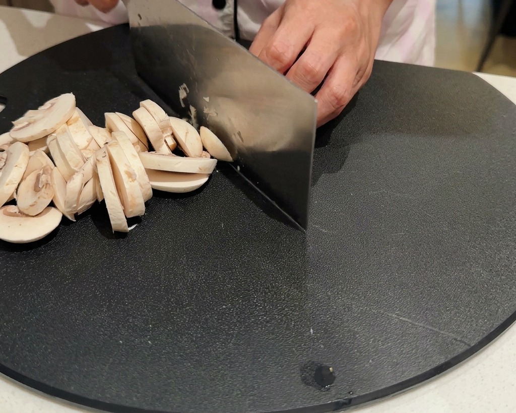 Hände schneiden mit einem großen Messer frische weiße Champignons auf einem schwarzen Schneidebrett in Scheiben.