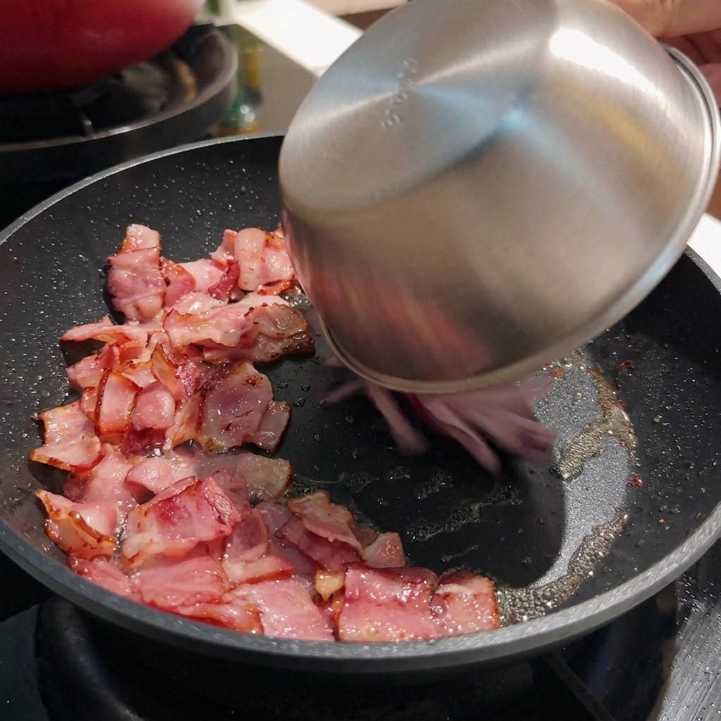 Geschnittene rote Zwiebeln werden aus einer Edelstahlschüssel in eine heiße Pfanne mit knusprigen Baconstücken gegeben.
