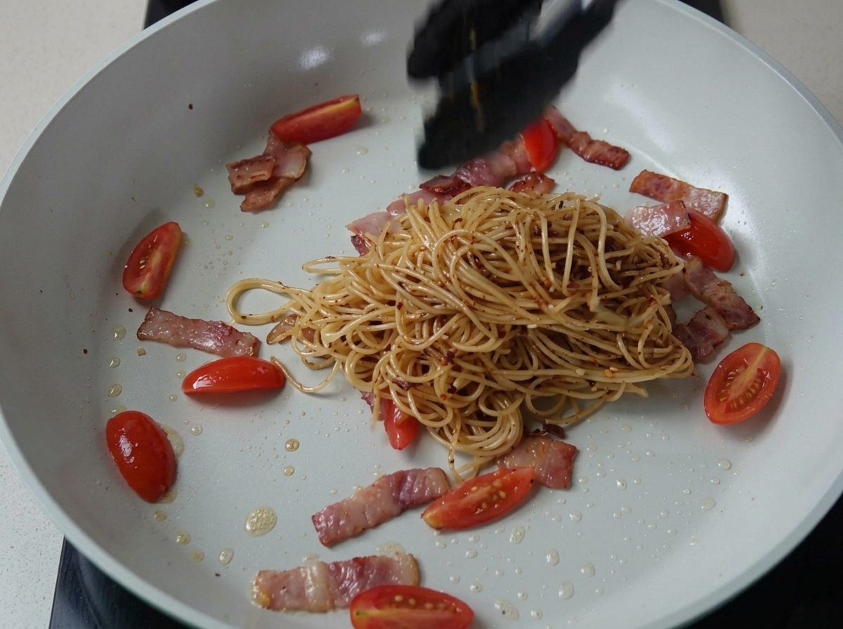 Gewürzte Spaghetti werden mit Speck und Tomaten in einer Pfanne geschwenkt.