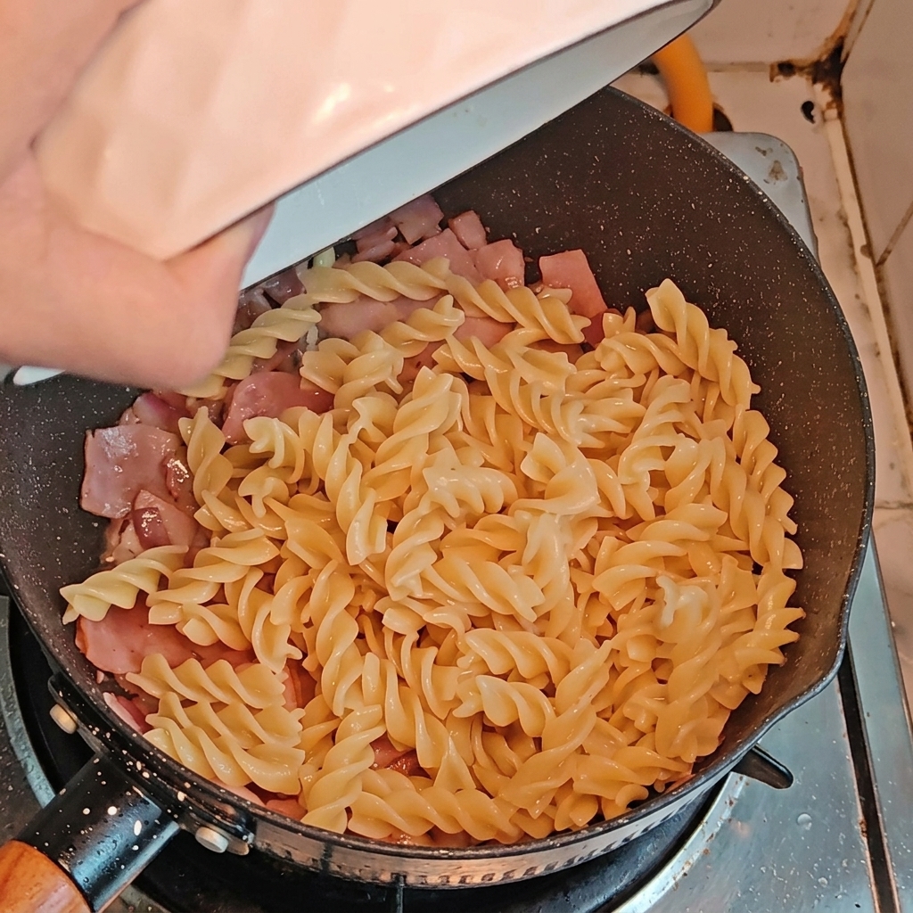 Gekochte Fusilli und geschnittener Spargel werden zu den gebratenen Zwiebeln und dem Speck in die Pfanne gegeben.