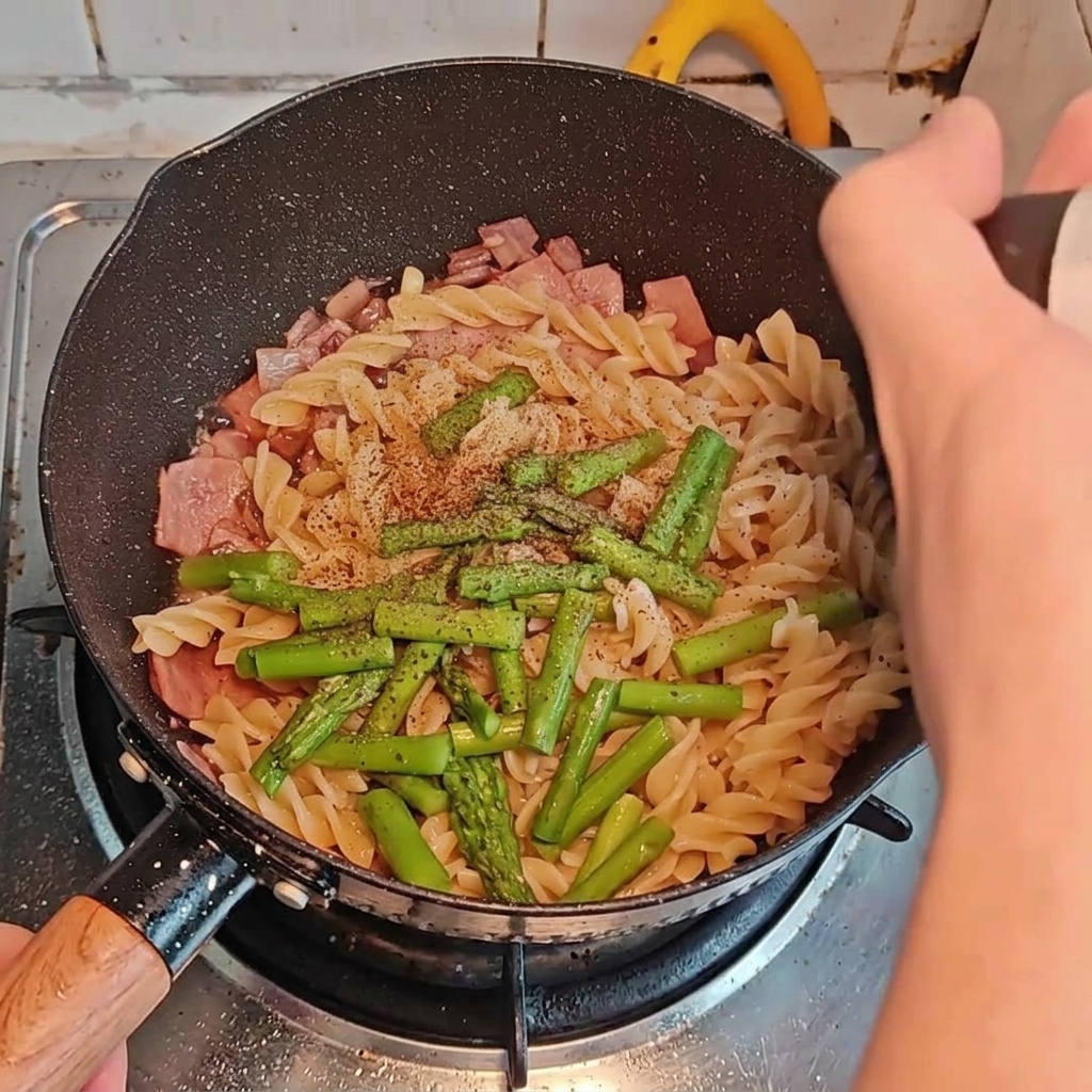 Verschiedene Gewürze werden über Pasta, Speck und Spargel in der Pfanne gestreut und gegossen.