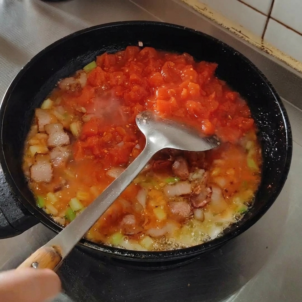 Ein Pfannenwender zerdrückt Tomaten in der Pfanne zu einer Sauce.