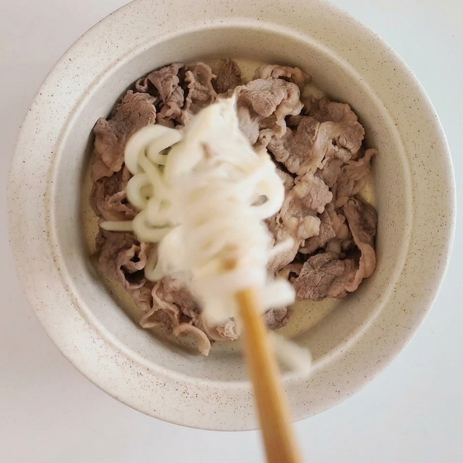 Eine angerichtete Schüssel mit zartem Rindfleisch, gekrönt von Udon-Nudeln und Enoki-Pilzen.