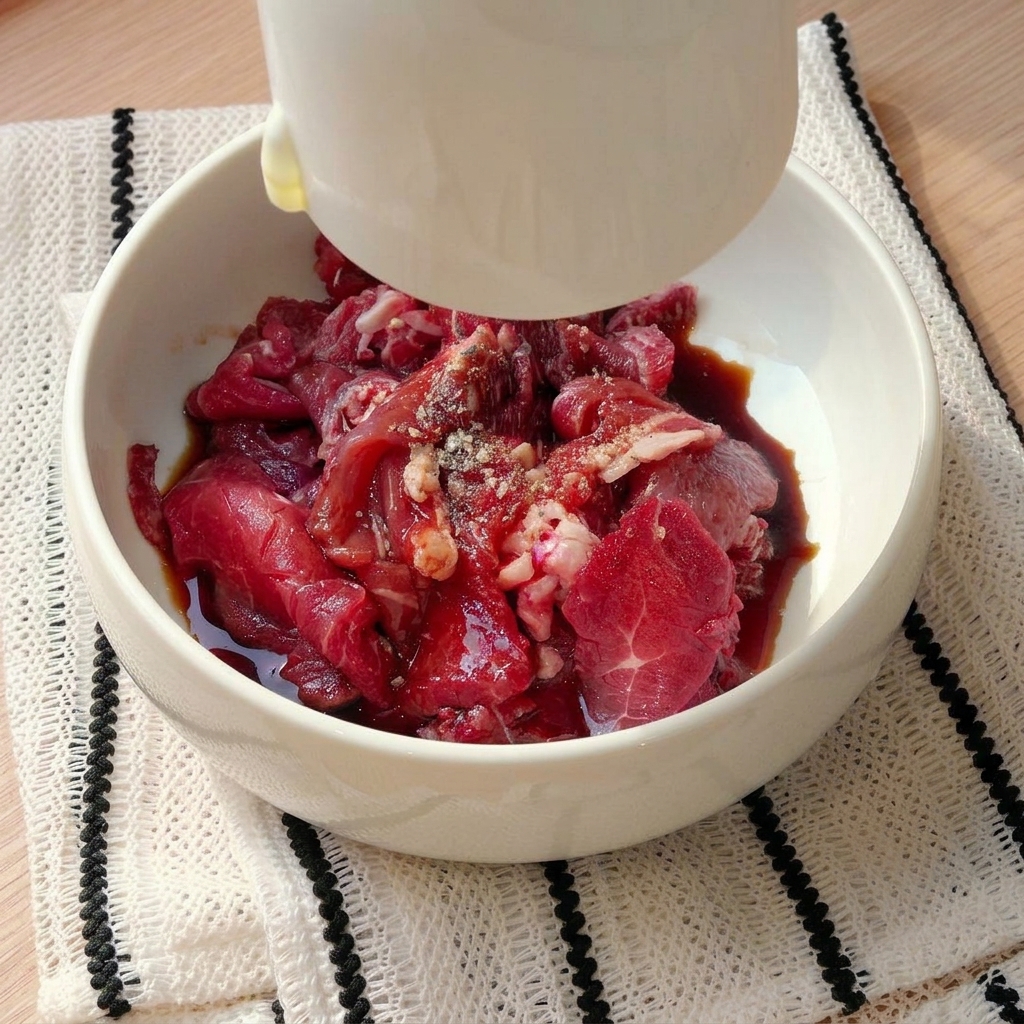 Rohe Rindfleischstreifen in einer weißen Schüssel, die mit flüssigen Zutaten und Gewürzen aus einer kleinen weißen Tasse gewürzt werden.