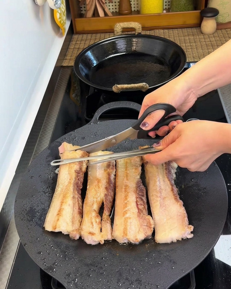 Eine Küchenschere schneidet die gebräunten Schweinebauchstreifen direkt in der Pfanne.