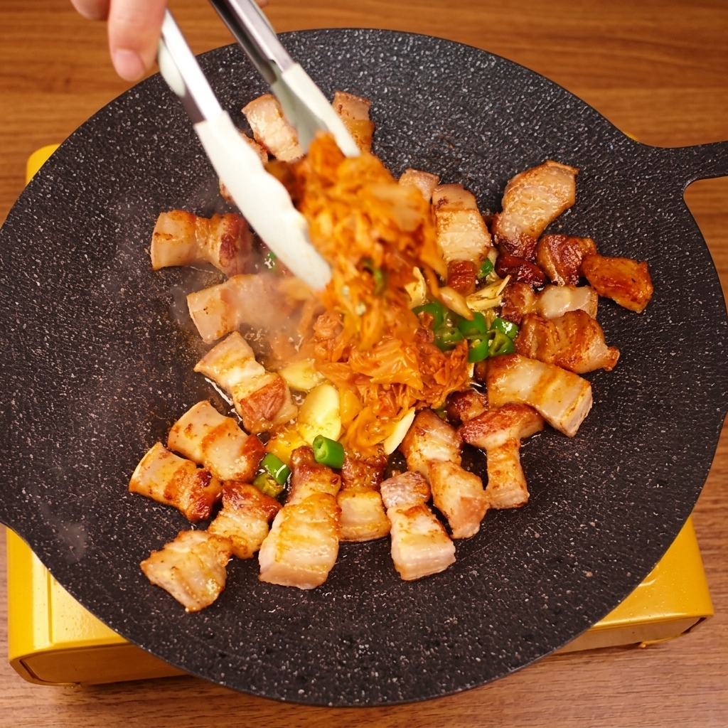 Mit einer Grillzange werden Schweinebauch und Kimchi in der Pfanne vermischt.