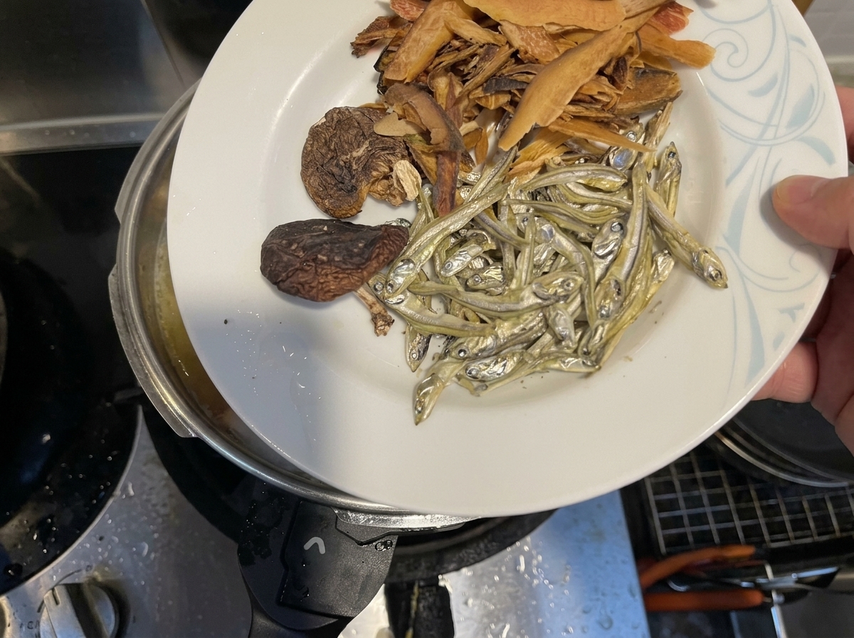 Trockenfisch, Shiitake-Pilze und Fischflocken werden in einen Topf mit köchelnder Flüssigkeit gegeben.