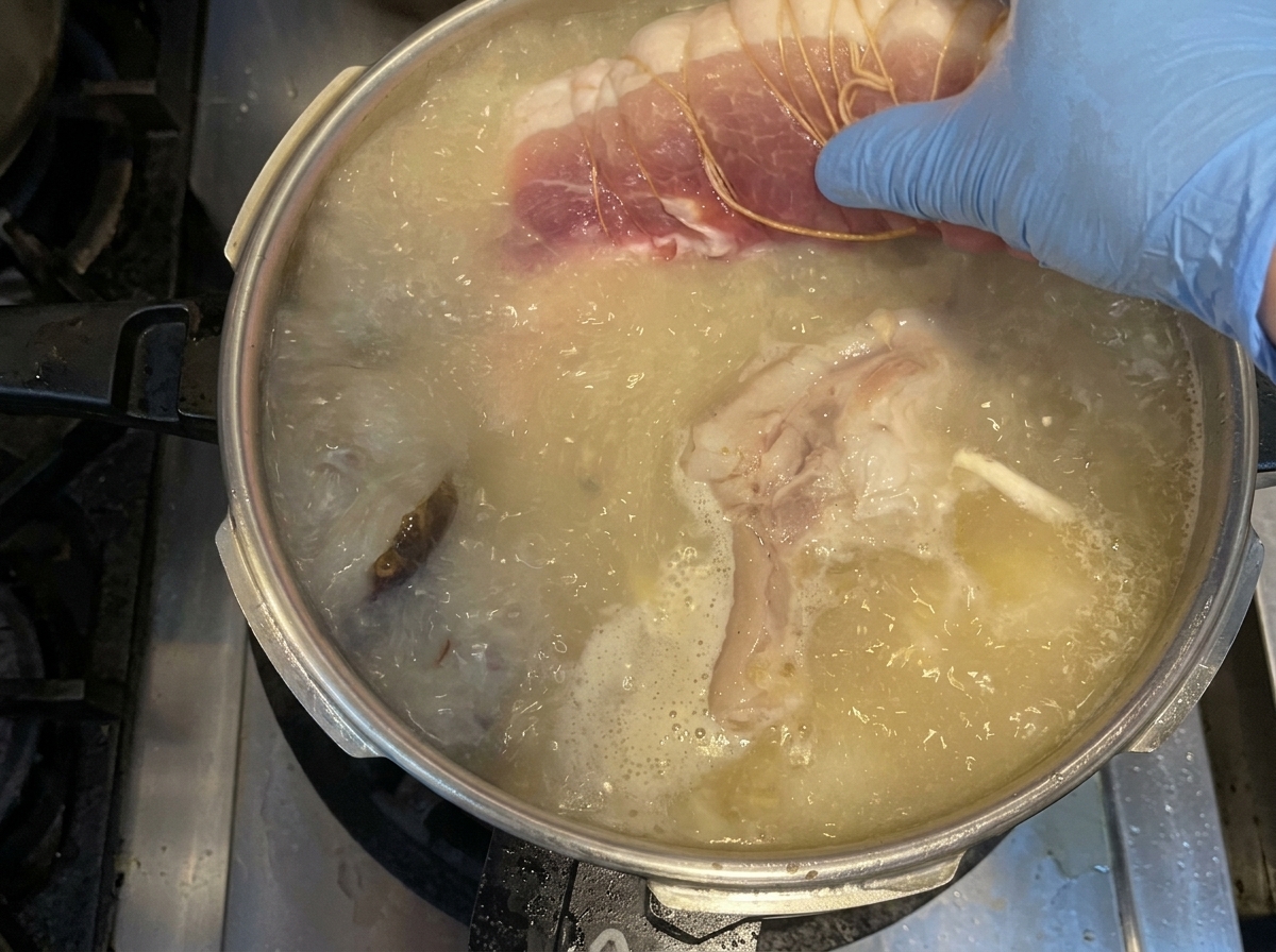 Eine Hand mit blauem Handschuh legt ein Stück rohe, gebundene Schweineschulter in einen Topf mit kochender Brühe und Gemüse.