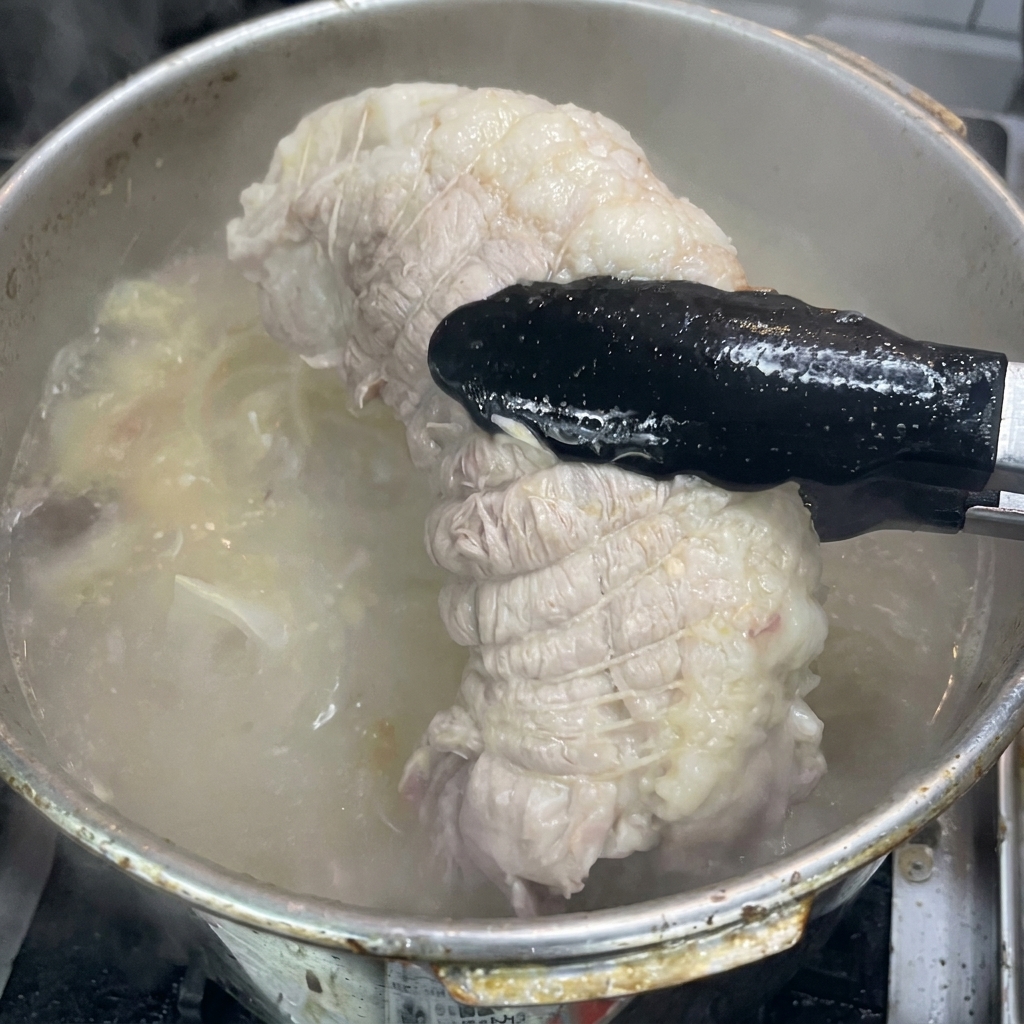 Eine schwarze Zange hebt ein fertig gekochtes, gebundenes Stück Schweineschulter aus einem Topf mit sprudelnder Brühe.