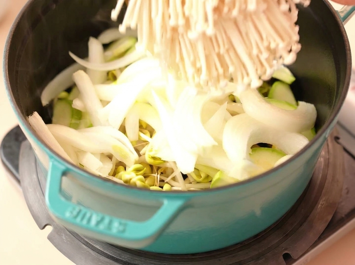 Ein Büschel Enoki-Pilze wird in einen Topf mit Zwiebeln, Zucchini und Sojasprossen gegeben.