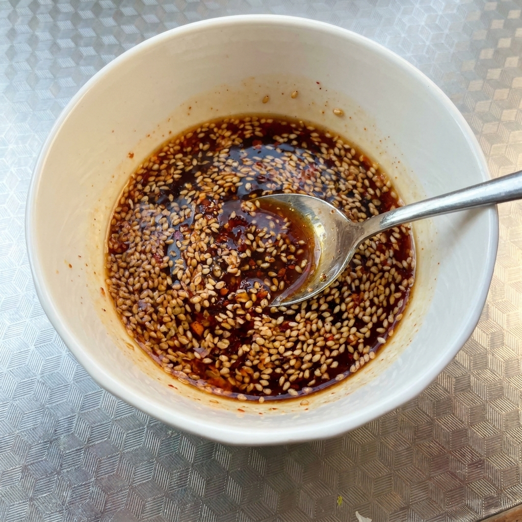 Eine kleine weiße Schüssel mit einer dunklen, würzigen Saucenmischung, die mit Sesam bestreut ist und mit einem Löffel verrührt wird.