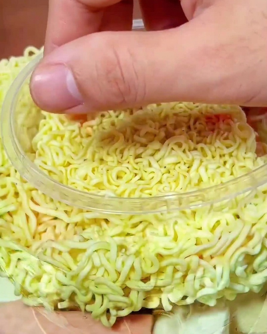 Eine Hand verteilt leuchtend rotes Kimchi über die Nudeln im Kochtopf.