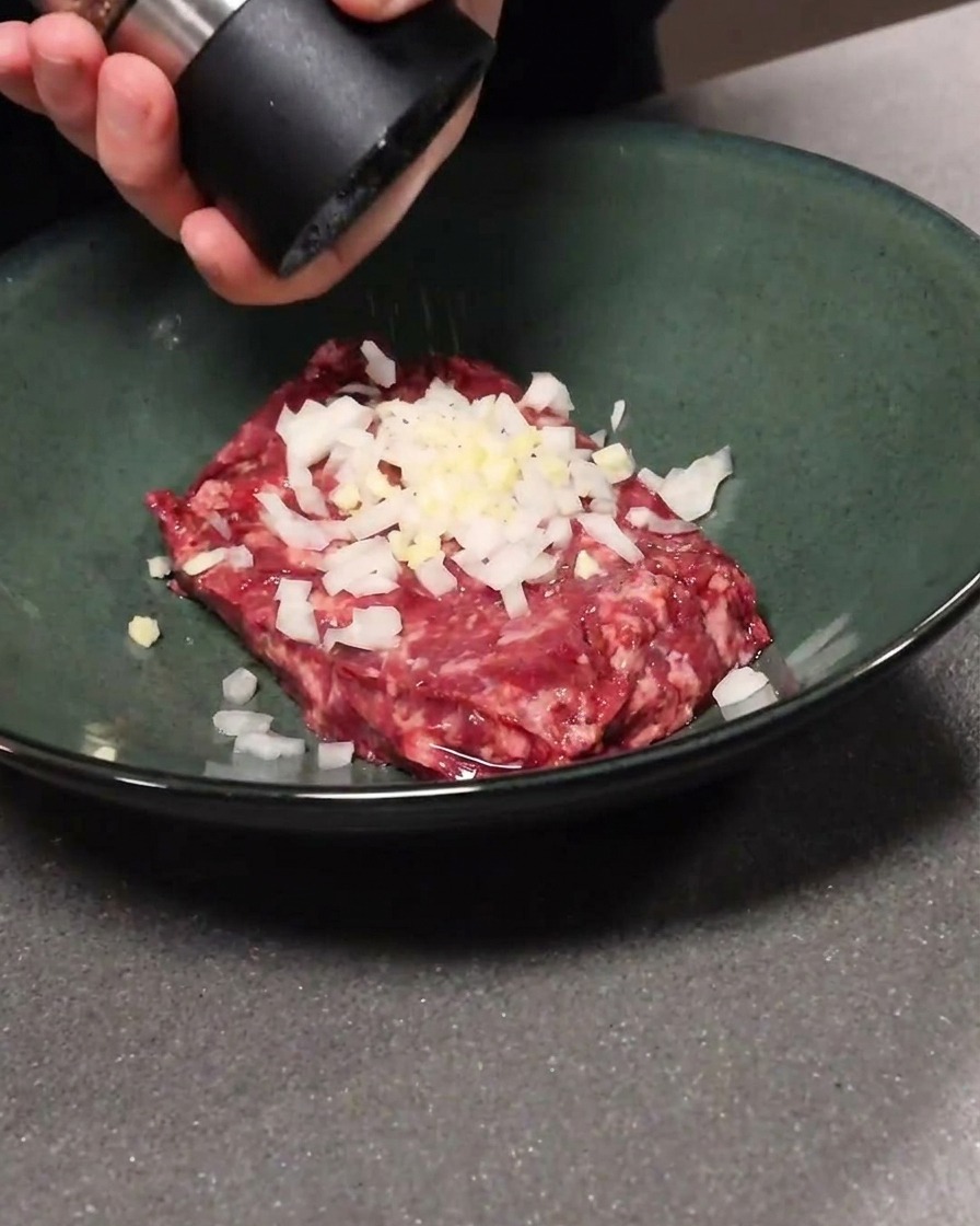 Rinderhackfleisch in einer Schüssel mit Knoblauch und Zwiebeln vermengt, während schwarzer Pfeffer hinzugefügt wird.