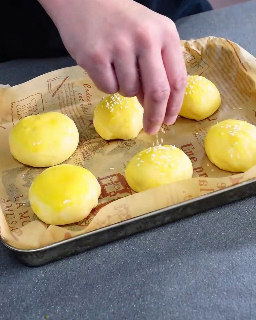 Weiße Sesamsamen werden über die mit Eistreiche bestrichenen, ungebackenen Brötchen gestreut.