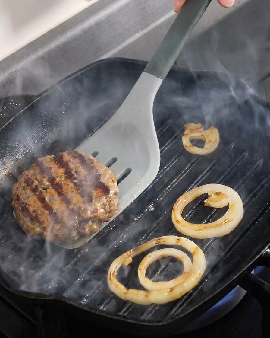 Nahaufnahme eines Rinder-Pattys und Zwiebelringen, die in einer heißen Grillpfanne gewendet werden.